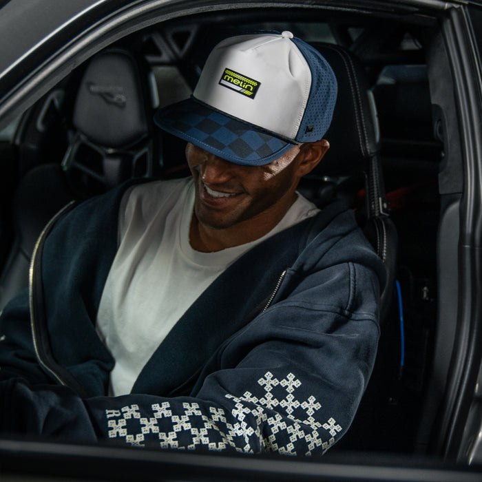 Black Iris and white colorblock trucker hat with embroidered checkerboard pattern on the bill, worn by man in car.
