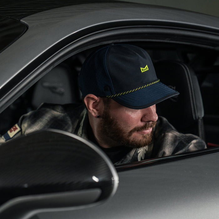 A man wearing the melin Split Fit Odysea Rope Bulls Icon MPH Hydro in  Black Iris / Black and a plaid shirt sits in the driver’s seat of a car, looking out the open window to his left.