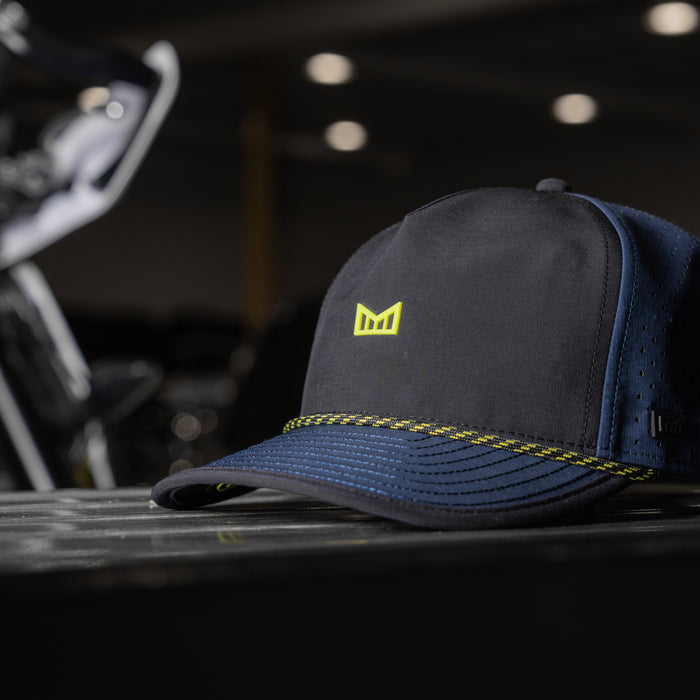 The melin Odysea MPH Hydro in Black Iris / Black rests on a surface, with a blurred background featuring bike handlebars.