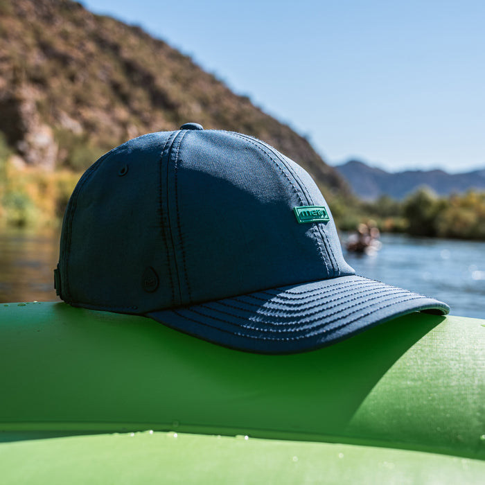 The melin Performance Dad Hat The Legend Hydro  in Deep Dive resting on an inflatable raft with water in the background