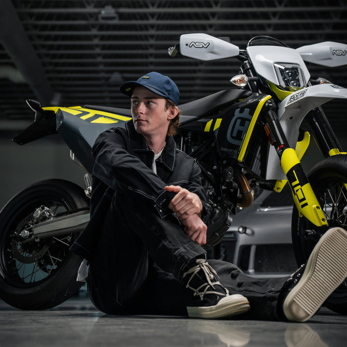 Man wearing the melin Performance Dad Hat The Legend MPH Hydro in  Black Iris / Black sitting on the ground next to a yellow and black Husqvarna motorcycle in a garage setting.