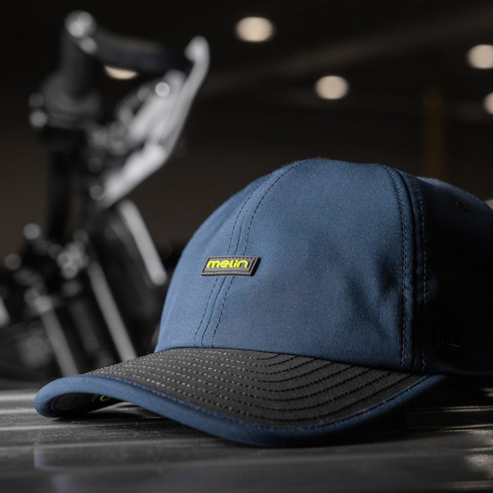 The melin Legend MPH Hydro in  Black Iris / Black rests on a striped surface, with a blurred background featuring bike handlebars.