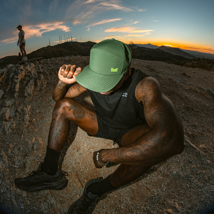 A man wearing the melin Trenches Icon Hydro in Kombu Green sits on a rocky hillside at sunset, resting his arm on his knee and tilting his head down.